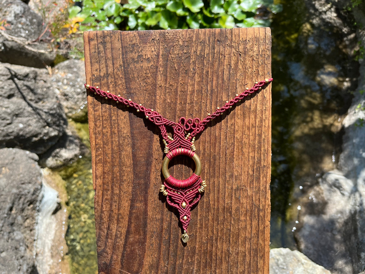 Burgundy macrame necklace with brass ring displayed on wood near water and rocks